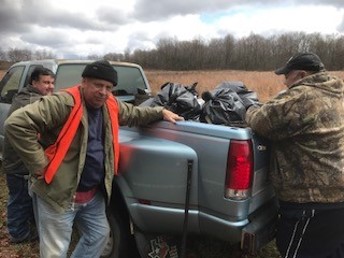 three men standing next to the back of a grey pickup truck. Black trash bags in the back of the pickup truck 